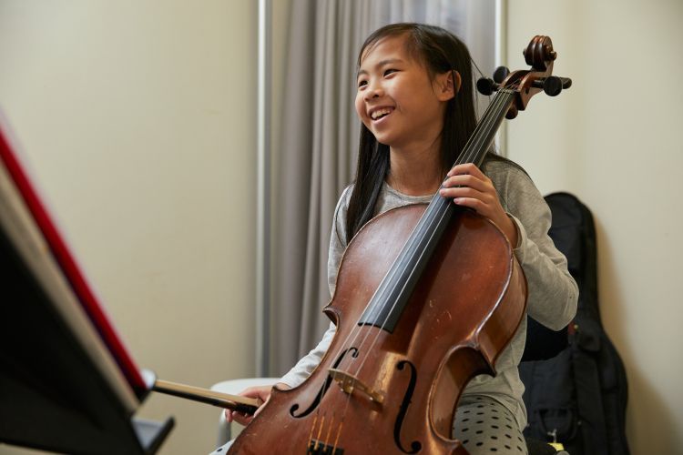 A girl with black hair and long sleeved grey top and trousers, sits at an angle holding a cello